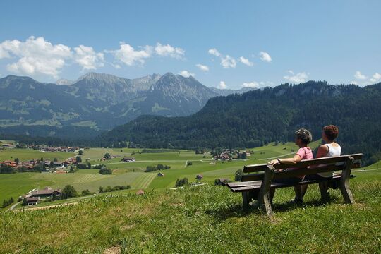 Two hikers sit on a bench high up on a beautiful summer's day and look down into the valley