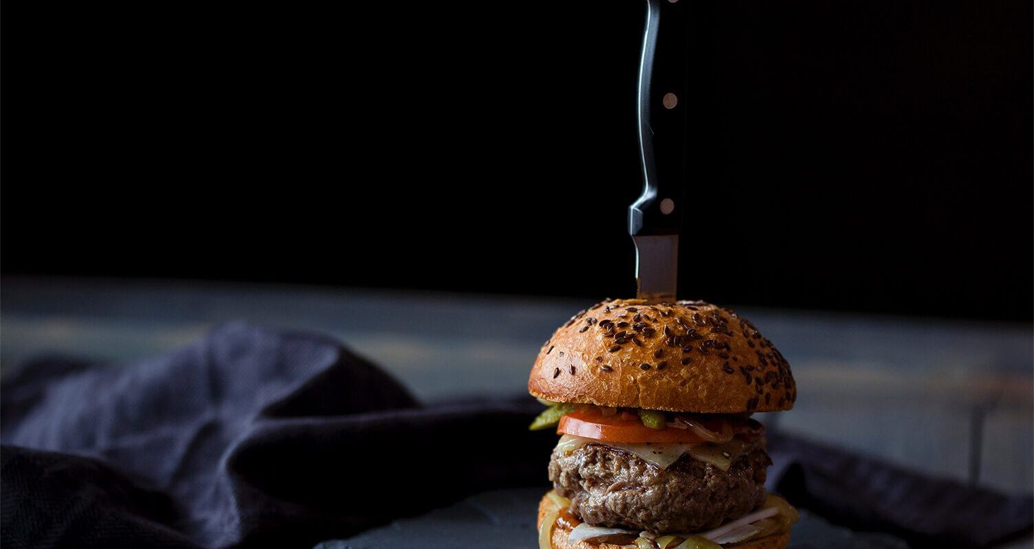 Ein Burger mit einem Kochmesser darin vor einem dunklen Hintergrund auf einer Schieferplatte