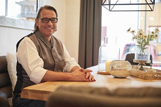 The hotel host sits at the regulars' table and smiles into the camera