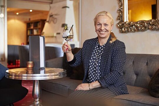 The hostess of the hotel sits on a bench with a summery drink from the bar and greets the camera