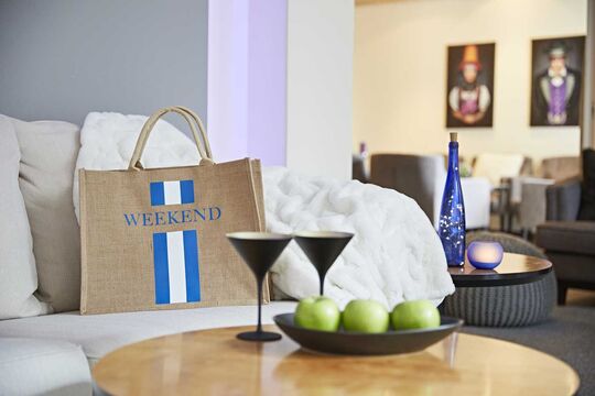 A bag labeled Weekend stands on a couch in the lobby of the hotel with a bowl of apples on a table in front of it