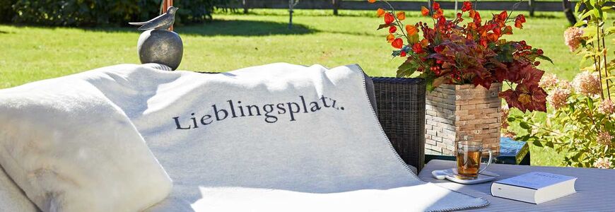 Eine Tagesdecke mit der Aufschrift Lieblingsplatz liegt über einer Outdoor Couch im Garten des Hotels an einem Sommertag