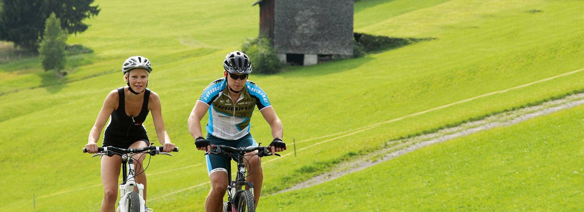Two cyclists with mountain bikes on a route on a mountain with a hut in the background