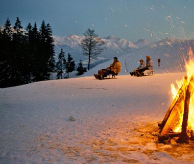 A campfire on a slope with tobogganers on sledges in the background in the early evening in winter